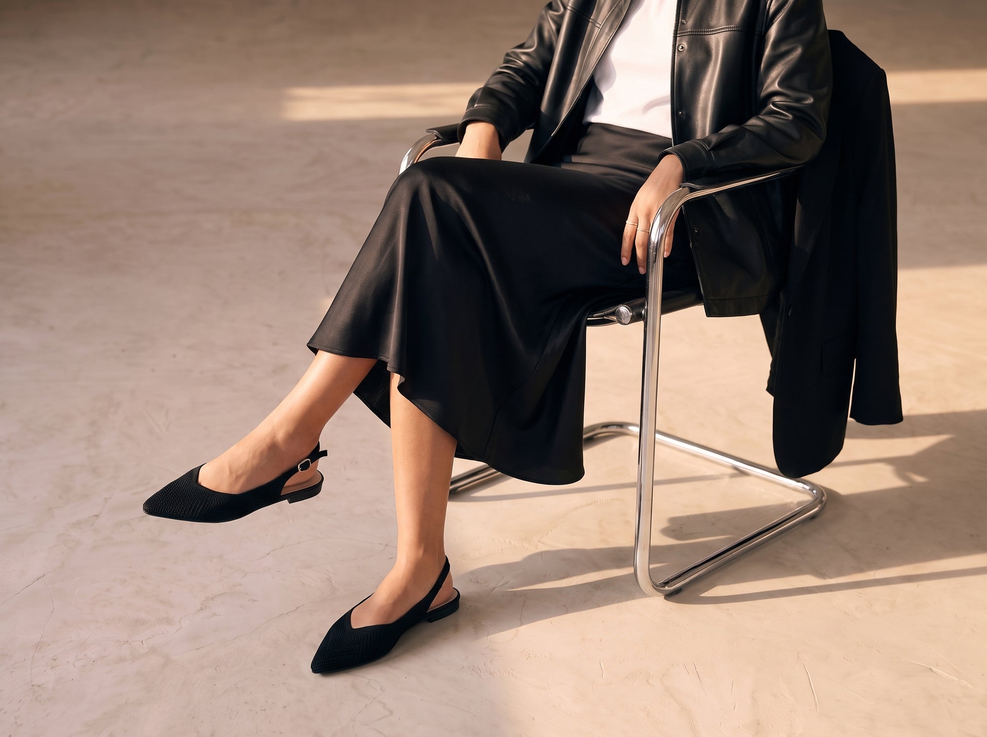 ALBA Minuit black knit slingback flats worn with a flowing black midi skirt while seated on a chrome chair.